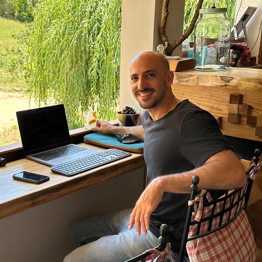 Eyal smiling at his laptop desk by a window with a willow tree outside