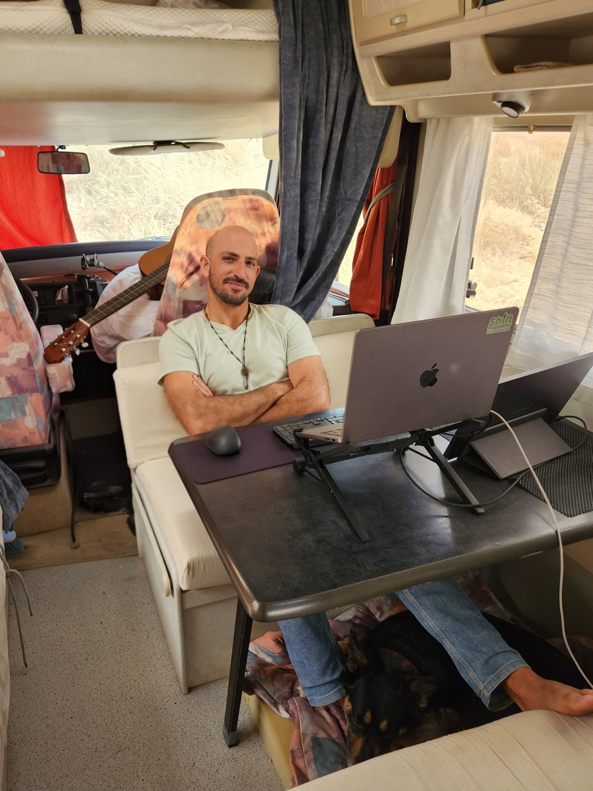 Eyal at the table inside his van with a laptop, a guitar behind him and his dog under the table